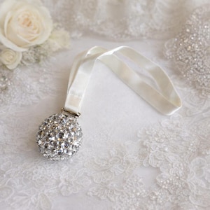 May include: A decorative bridal garter featuring a sparkling, crystal-covered ball and a cream-colored velvet ribbon. The garter is designed with a silver-tone metal clasp and rests on a white lace surface, with white roses in the background.