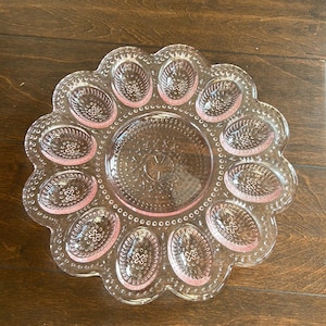 May include: A clear, pink glass deviled egg serving platter with a scalloped edge. The platter has 12 oval-shaped indentations around the edge and a circular center. The glass has a textured design.