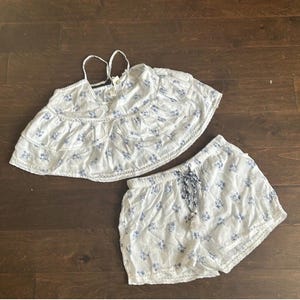 May include: A white two-piece set featuring a sleeveless tiered crop top with thin spaghetti straps and matching shorts. Both pieces are decorated with a small blue floral pattern and delicate lace trim details. The shorts include a drawstring waist.