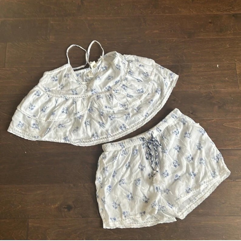 May include: A white two-piece set featuring a sleeveless tiered crop top with thin spaghetti straps and matching shorts. Both pieces are decorated with a small blue floral pattern and delicate lace trim details. The shorts include a drawstring waist.