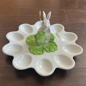 May include: A white ceramic egg holder with a scalloped edge, featuring a decorative bunny figurine atop a green lettuce-shaped base. The egg tray has 10 egg-shaped indentations. A charming Easter-themed serving piece.
