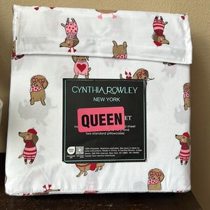 May include: White queen-size sheet set with a repeating pattern of dachshund dogs wearing sweaters, hats, and glasses. The set includes a fitted sheet and two standard pillowcases. The packaging features the Cynthia Rowley New York logo.