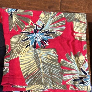 Puede incluir: Una pieza de tela doblada con un fondo rojo vibrante y un patr&oacute;n tropical repetitivo de grandes hojas de palmera de color verde claro y flores de color azul oscuro en forma de estrella. La tela tiene un borde dobladillado con costuras rojas.