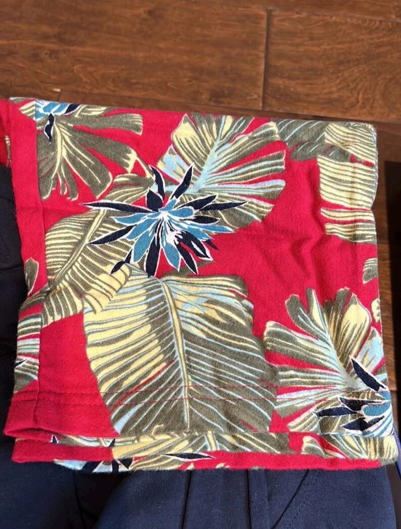Puede incluir: Una pieza de tela doblada con un fondo rojo vibrante y un patr&oacute;n tropical repetitivo de grandes hojas de palmera de color verde claro y flores de color azul oscuro en forma de estrella. La tela tiene un borde dobladillado con costuras rojas.