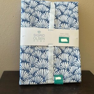 May include: A folded tablecloth with a blue and white fan-shaped pattern. The label reads "SIGRID OLSEN HOME" and "EASY CARE TABLECLOTH". A white ribbon wraps around the folded tablecloth.