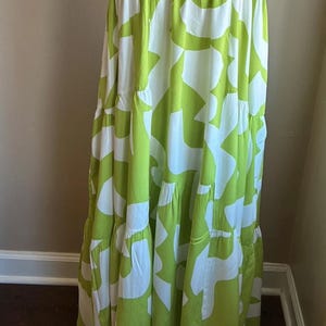 May include: A floor-length, sleeveless dress with a white and lime green abstract pattern. The dress features a square neckline and wide straps. The fabric is lightweight and flowy, with tiered ruffles at the skirt.