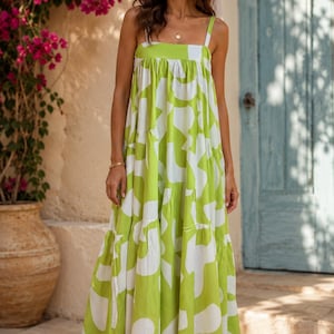 May include: A long, flowing dress in a vibrant lime green and white abstract pattern. The dress features a square neckline, wide straps, and a tiered skirt. The model is wearing brown sandals and a gold necklace.
