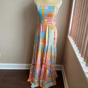 Pode incluir: Um vestido maxi sem al&ccedil;as com um design de patchwork. O vestido apresenta uma mistura de cores, incluindo azul, laranja, rosa e amarelo. O corpete &eacute; franzido e a saia tem babados em camadas. O vestido &eacute; exibido em um manequim.