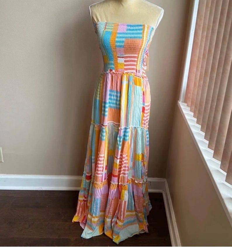 Pode incluir: Um vestido maxi sem al&ccedil;as com um design de patchwork. O vestido apresenta uma mistura de cores, incluindo azul, laranja, rosa e amarelo. O corpete &eacute; franzido e a saia tem babados em camadas. O vestido &eacute; exibido em um manequim.