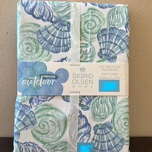May include: A folded tablecloth featuring a seashell pattern in shades of blue and green on a white background. The tablecloth is wrapped with a ribbon and has a label that reads "Sigrid Olsen Home" and "Indoor Outdoor".