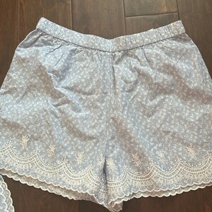 May include: Light blue shorts featuring a white floral pattern and an elastic waistband. The hem is finished with a wide, white scalloped embroidery detail that includes intricate floral stitching along the bottom edge of the garment.