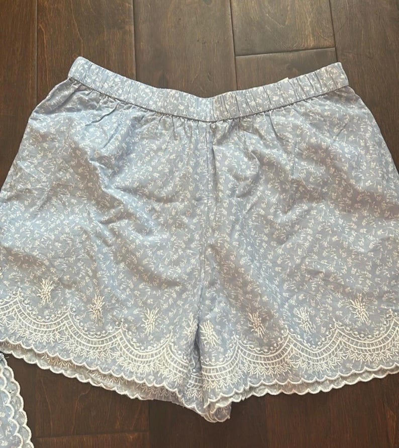 May include: Light blue shorts featuring a white floral pattern and an elastic waistband. The hem is finished with a wide, white scalloped embroidery detail that includes intricate floral stitching along the bottom edge of the garment.