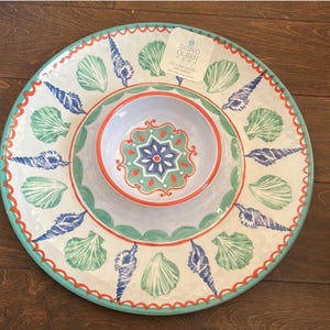 May include: A round, decorative serving platter and bowl set. The set features a white background with blue, green, and orange seashell designs. The bowl has a floral pattern in the center. The rim is scalloped with a turquoise border.