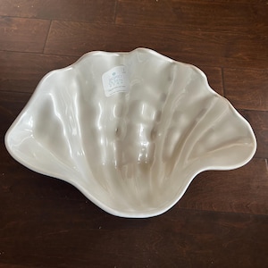May include: A large, off-white ceramic seashell-shaped bowl. The bowl has a glossy finish and a textured surface, with a label that reads "SIGRID OLSEN HOME". The bowl is placed on a dark brown wooden surface.