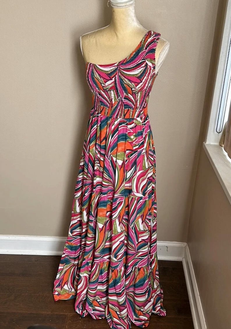 May include: A colorful, one-shoulder maxi dress with a vibrant leaf-like pattern in shades of pink, orange, green, and white. The dress features a fitted bodice and a flowing, tiered skirt, perfect for warm weather.