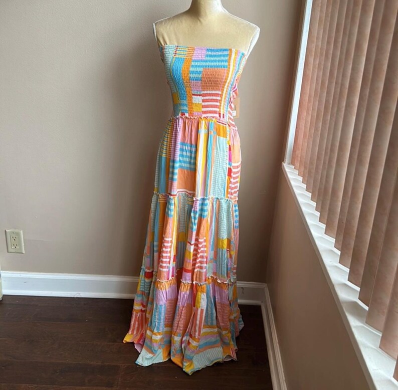 May include: A strapless maxi dress in a patchwork design. The dress features a mix of colors including blue, orange, pink, and yellow. The bodice is smocked, and the skirt has tiered ruffles. The dress is displayed on a mannequin.