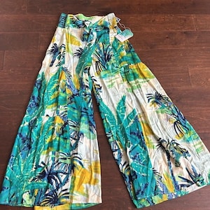 May include: Wide-leg pants with a tropical print featuring green, blue, and yellow palm leaves on a white background. The pants have a drawstring waist and a tag attached. The fabric appears lightweight and suitable for warm weather.