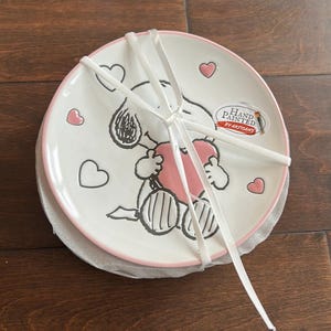 May include: Set of two white ceramic plates with a pink rim, featuring a hand-painted Snoopy design holding a pink heart. The plates are tied together with a white ribbon and include heart accents. The plates are labeled "Hand Painted by Artisans."