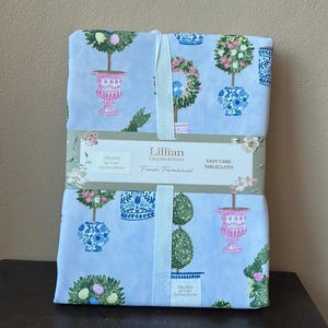May include: Oblong tablecloth featuring a light blue background with a repeating pattern of potted topiaries in pink, blue, and green. The tablecloth is tied with a white ribbon and includes the text "Lillian August French Farmhouse". The dimensions are 60 x 104 inches.