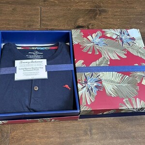 Puede incluir: Una camiseta henley azul marino de manga larga con un peque&ntilde;o logotipo de marl&iacute;n rojo y un bot&oacute;n, empaquetada en una caja con tapa estampada de hojas tropicales rojas y verdes. Una etiqueta blanca dice Tommy Bahama Long Sleeve Henley Top & Flannel Pant.