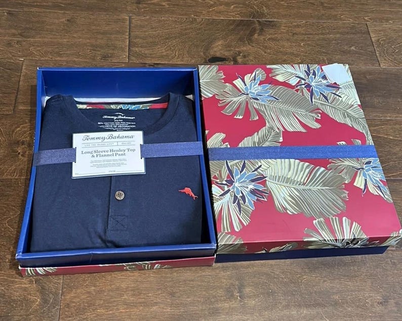Puede incluir: Una camiseta henley azul marino de manga larga con un peque&ntilde;o logotipo de marl&iacute;n rojo y un bot&oacute;n, empaquetada en una caja con tapa estampada de hojas tropicales rojas y verdes. Una etiqueta blanca dice Tommy Bahama Long Sleeve Henley Top & Flannel Pant.