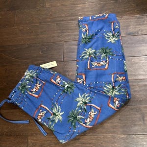 Puede incluir: Pantalones de pijama azules con un estampado tropical con palmeras, figuras en hamacas y luces de cadena. Los pantalones tienen cintura con cord&oacute;n y una etiqueta adjunta. La tela parece suave y ligera.