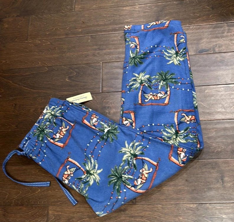 Puede incluir: Pantalones de pijama azules con un estampado tropical con palmeras, figuras en hamacas y luces de cadena. Los pantalones tienen cintura con cord&oacute;n y una etiqueta adjunta. La tela parece suave y ligera.