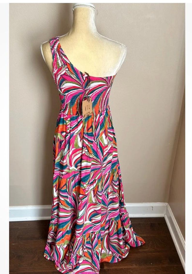 May include: A colorful, one-shoulder maxi dress with a vibrant abstract pattern in pink, orange, green, and white. The dress features a fitted bodice and a flowing skirt. A tag with the letters "ITL" is attached.