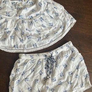 May include: A two-piece set consisting of a white sleeveless top and matching shorts, both featuring a blue floral print and vertical stripes. The top has tiered ruffled layers with lace trim, while the shorts include a drawstring waist with tassels.