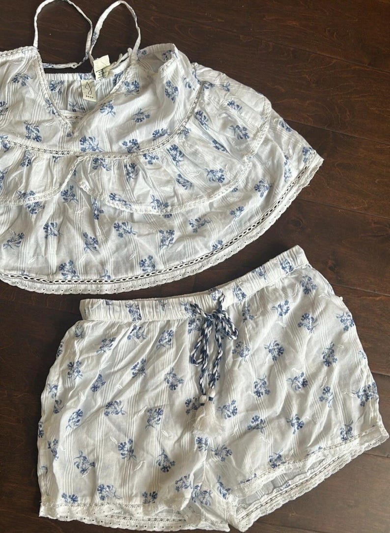 May include: A two-piece set consisting of a white sleeveless top and matching shorts, both featuring a blue floral print and vertical stripes. The top has tiered ruffled layers with lace trim, while the shorts include a drawstring waist with tassels.