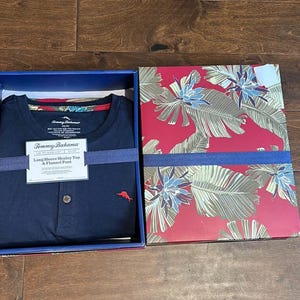 Puede incluir: Una camiseta henley de manga larga en color azul marino con un peque&ntilde;o logotipo de pez bordado en rojo en el pecho, presentada en una caja con tapa roja que muestra un estampado de hojas y flores tropicales. Etiqueta con Tommy Bahama.