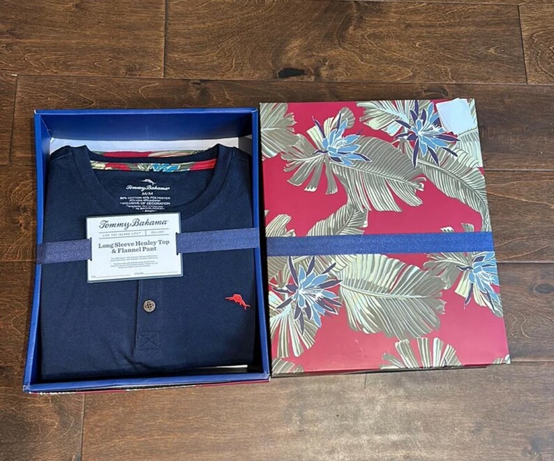 Puede incluir: Una camiseta henley de manga larga en color azul marino con un peque&ntilde;o logotipo de pez bordado en rojo en el pecho, presentada en una caja con tapa roja que muestra un estampado de hojas y flores tropicales. Etiqueta con Tommy Bahama.