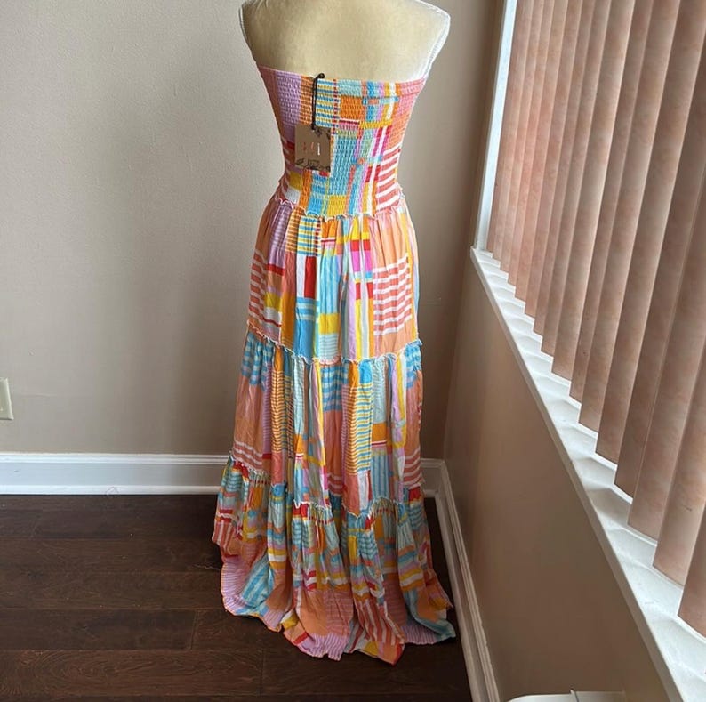 May include: A colorful strapless maxi dress with a patchwork design. The dress features a mix of orange, pink, blue, and yellow rectangular patterns. The dress has a tiered skirt and is displayed on a mannequin.
