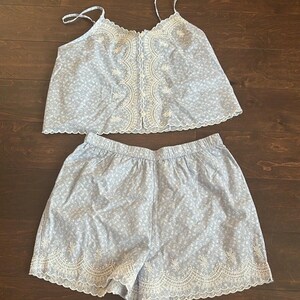 May include: A light blue two-piece set consisting of a camisole and shorts. The camisole features thin straps, a button-down front, and white floral embroidery along the neckline and placket. The shorts have an elastic waistband and white floral embroidery along the scalloped hem.