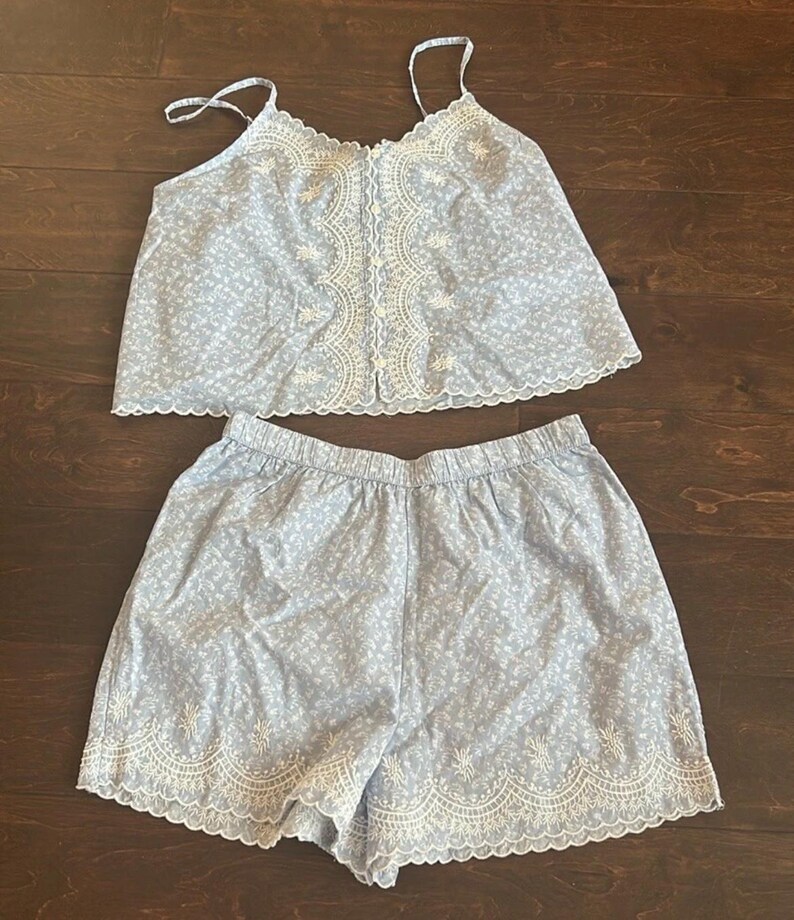 May include: A light blue two-piece set consisting of a camisole and shorts. The camisole features thin straps, a button-down front, and white floral embroidery along the neckline and placket. The shorts have an elastic waistband and white floral embroidery along the scalloped hem.
