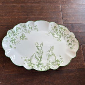 May include: White oval serving platter with a scalloped edge and green floral and bunny design. The platter features two bunnies and floral accents in a green hue. The rim is also green.