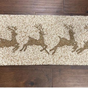 May include: A rectangular table runner with a cream-colored background and a pattern of four running reindeer in a gold color. The runner is placed on a dark brown wooden surface.
