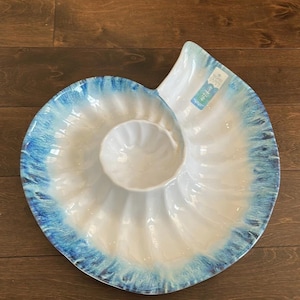May include: White ceramic serving platter in a nautilus shell design, with a central dip bowl. The rim features a blue, textured, watercolor-style design. A decorative piece for serving appetizers or snacks.