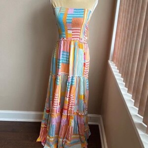Pode incluir: Um vestido maxi sem al&ccedil;as com um design de patchwork colorido. O vestido apresenta um corpete franzido e uma saia em camadas. A paleta de cores inclui tons de azul, laranja, rosa e amarelo. O vestido &eacute; exibido em um manequim.