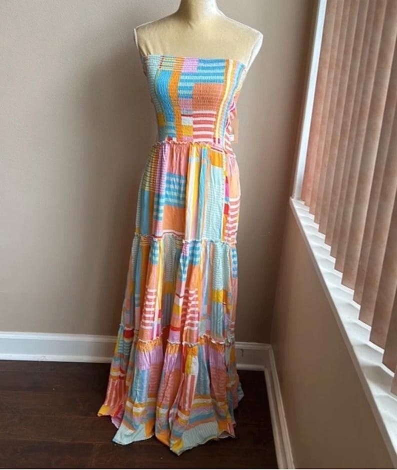 Pode incluir: Um vestido maxi sem al&ccedil;as com um design de patchwork colorido. O vestido apresenta um corpete franzido e uma saia em camadas. A paleta de cores inclui tons de azul, laranja, rosa e amarelo. O vestido &eacute; exibido em um manequim.