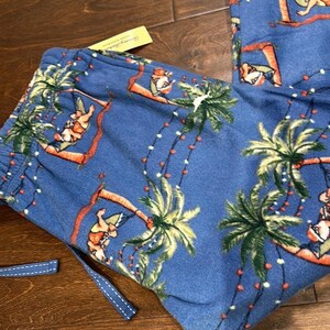 Puede incluir: Pantalones de pijama azules con un tema navide&ntilde;o tropical. Los pantalones presentan un patr&oacute;n repetido de palmeras, figuras de Pap&aacute; Noel y luces de cadena. El cord&oacute;n es azul con costuras blancas. Se adjunta una etiqueta amarilla.