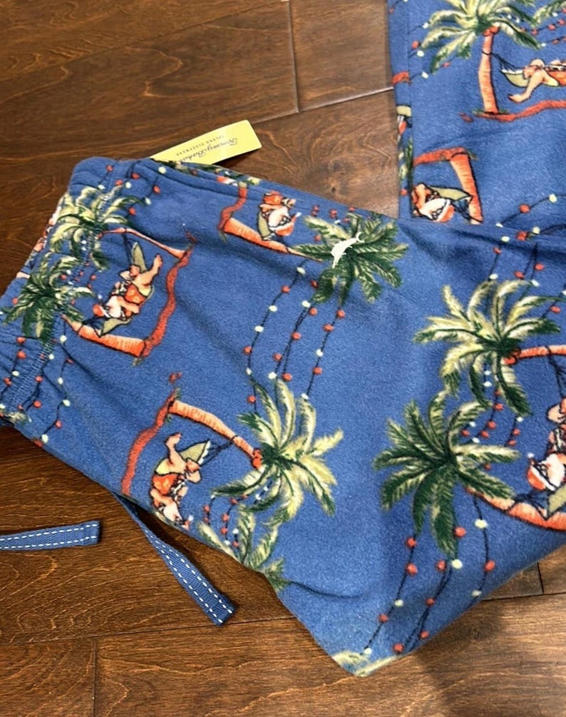 Puede incluir: Pantalones de pijama azules con un tema navide&ntilde;o tropical. Los pantalones presentan un patr&oacute;n repetido de palmeras, figuras de Pap&aacute; Noel y luces de cadena. El cord&oacute;n es azul con costuras blancas. Se adjunta una etiqueta amarilla.