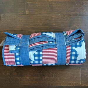 May include: Rolled-up picnic blanket featuring a patriotic design. The blanket displays red and white stripes, blue gingham, and white stars. It is secured with blue fabric straps. The blanket is rolled up on a dark wood surface.