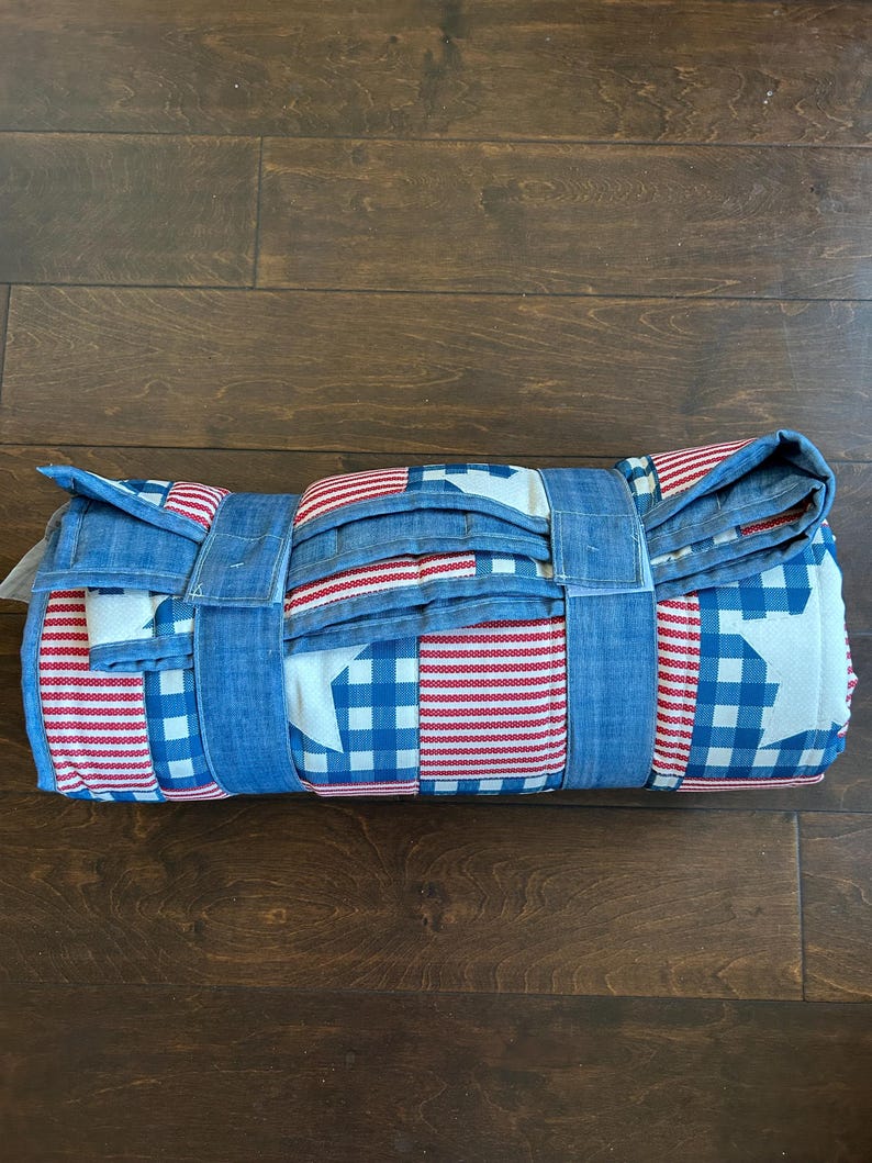 May include: Rolled-up picnic blanket featuring a patriotic design. The blanket displays red and white stripes, blue gingham, and white stars. It is secured with blue fabric straps. The blanket is rolled up on a dark wood surface.