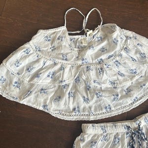 May include: A white sleeveless top with a blue floral print, featuring two horizontal ruffled tiers with white eyelet lace trim, thin spaghetti straps, and a V-neckline. The fabric has a subtle vertical texture and a small brand tag at the neck.