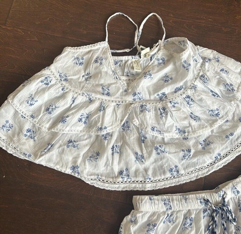 May include: A white sleeveless top with a blue floral print, featuring two horizontal ruffled tiers with white eyelet lace trim, thin spaghetti straps, and a V-neckline. The fabric has a subtle vertical texture and a small brand tag at the neck.