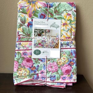 May include: A folded floral tablecloth with a tag that reads "Eternal Rose Tablecloth." The tablecloth features a pattern of pink roses, blue and yellow flowers, and green leaves on a white background. It is tied with a pink and white string.