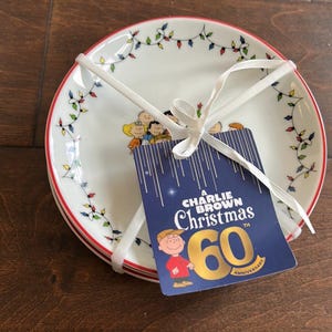 May include: Set of three white ceramic plates with a red rim, decorated with colorful Christmas lights and Peanuts characters. A blue tag tied with a white ribbon reads "A Charlie Brown Christmas 60th Anniversary."