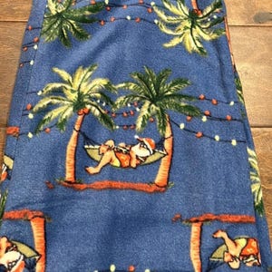 Puede incluir: Pantalones de pijama azules con un estampado repetido de palmeras, Pap&aacute; Noel en una hamaca y luces de cadena. Los pantalones est&aacute;n hechos de un material suave.