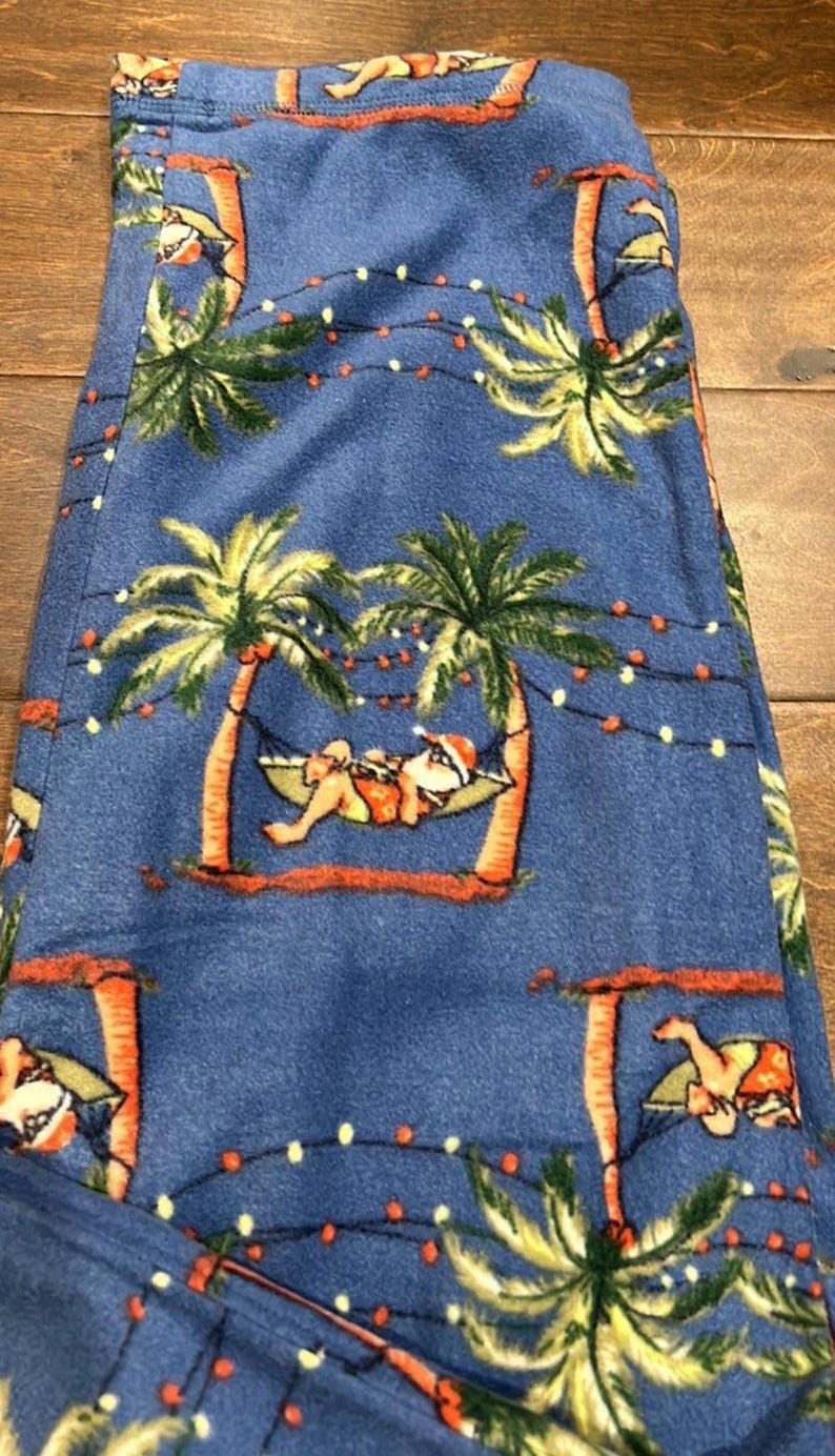 Puede incluir: Pantalones de pijama azules con un estampado repetido de palmeras, Pap&aacute; Noel en una hamaca y luces de cadena. Los pantalones est&aacute;n hechos de un material suave.